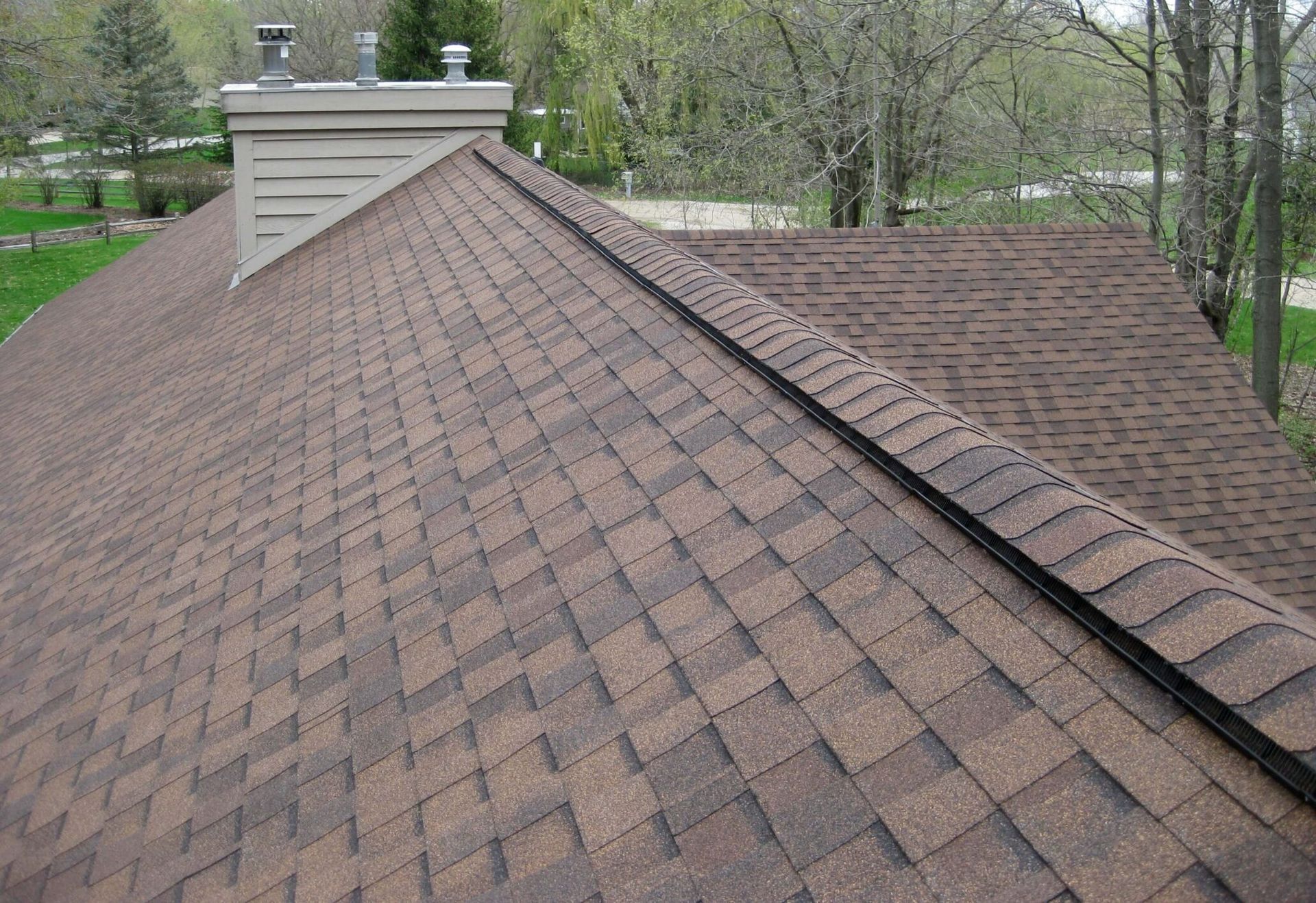 Reddish-brown shingle roof with a central ridge vent and a chimney on a wooded home.