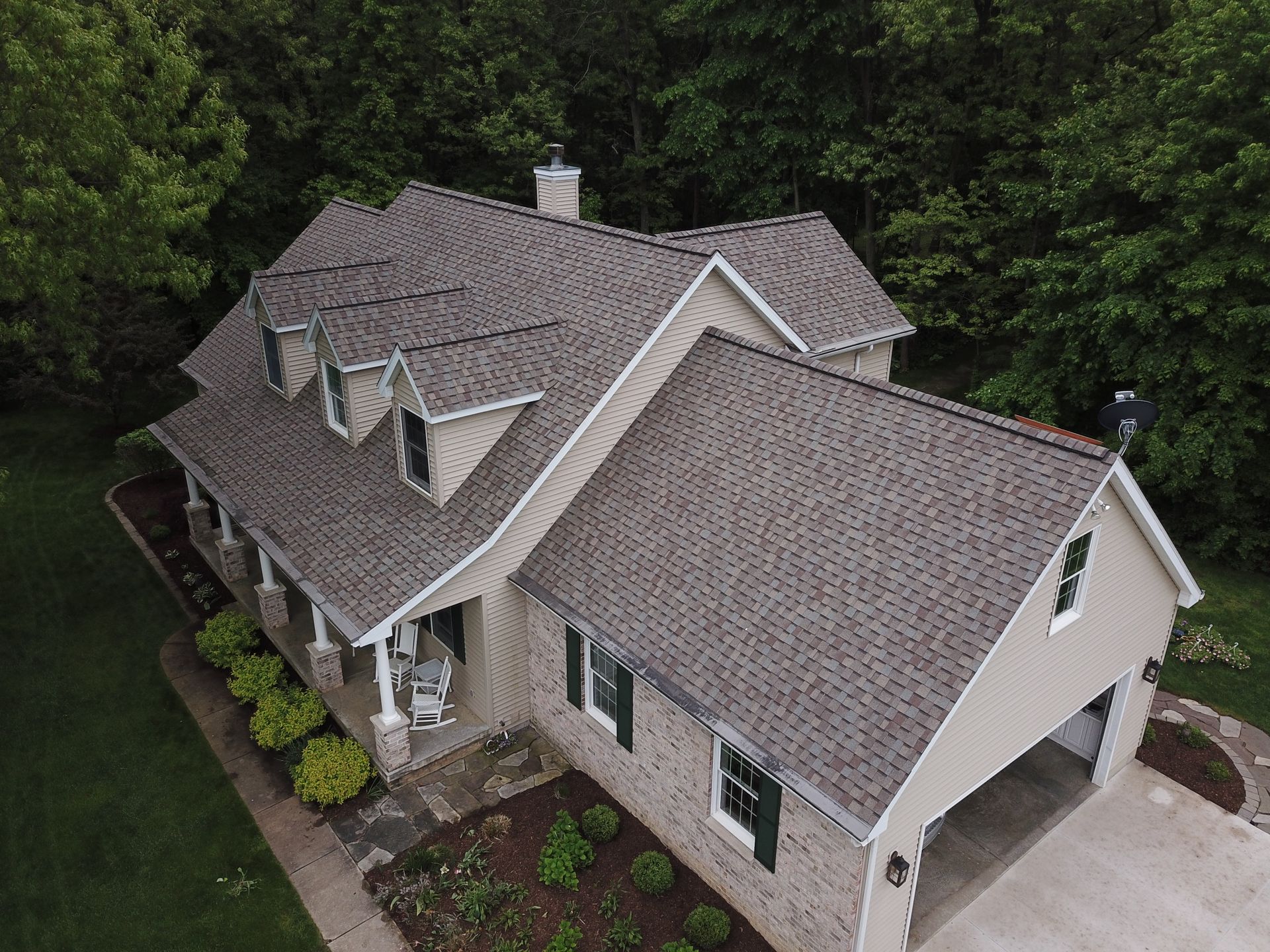Aerial view of a beige house with gray shingle roof beside a driveway, surrounded by trees