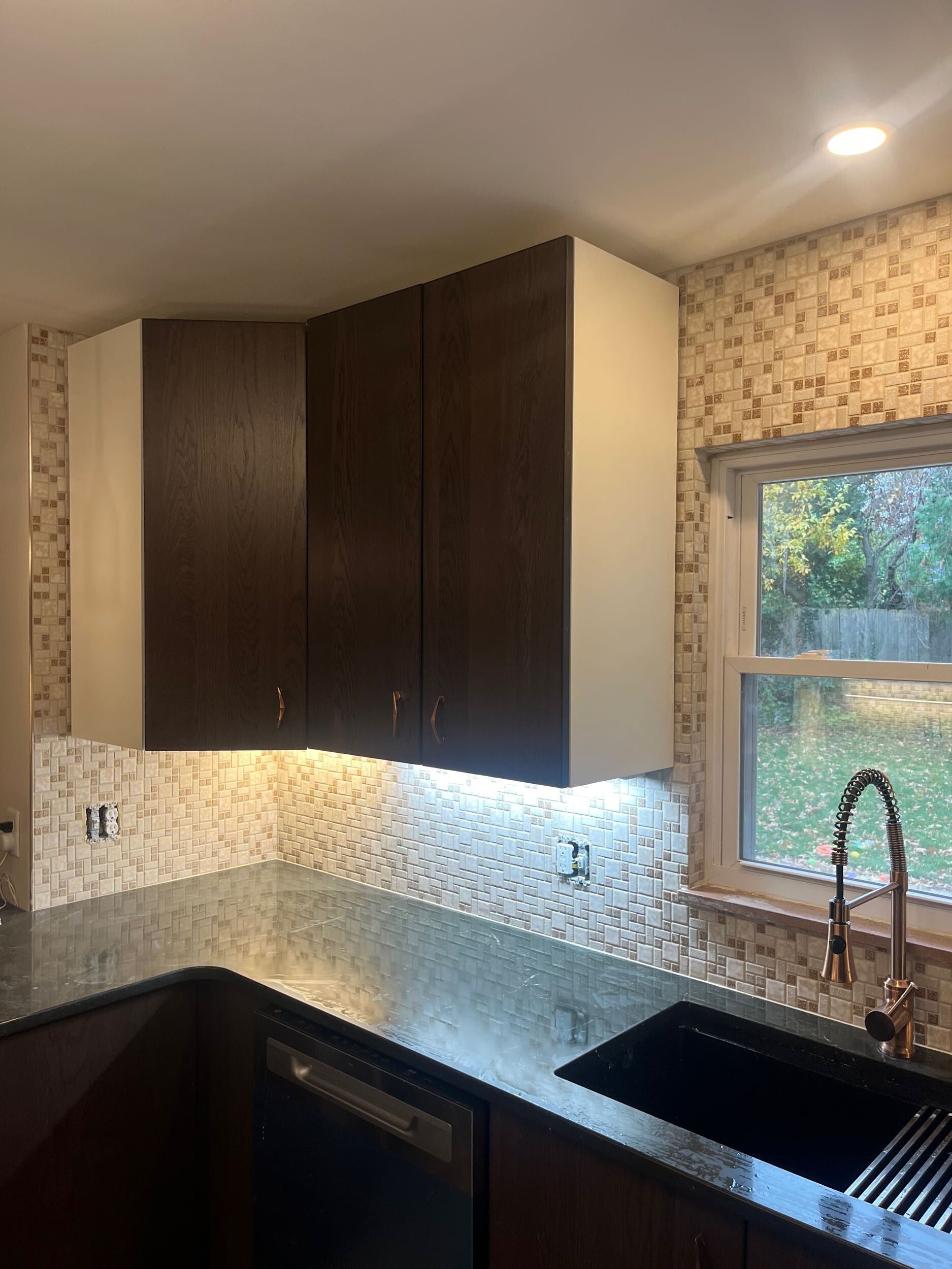 Modern kitchen corner with dark cabinets, granite counters, tiled backsplash, sink, and window above the sink