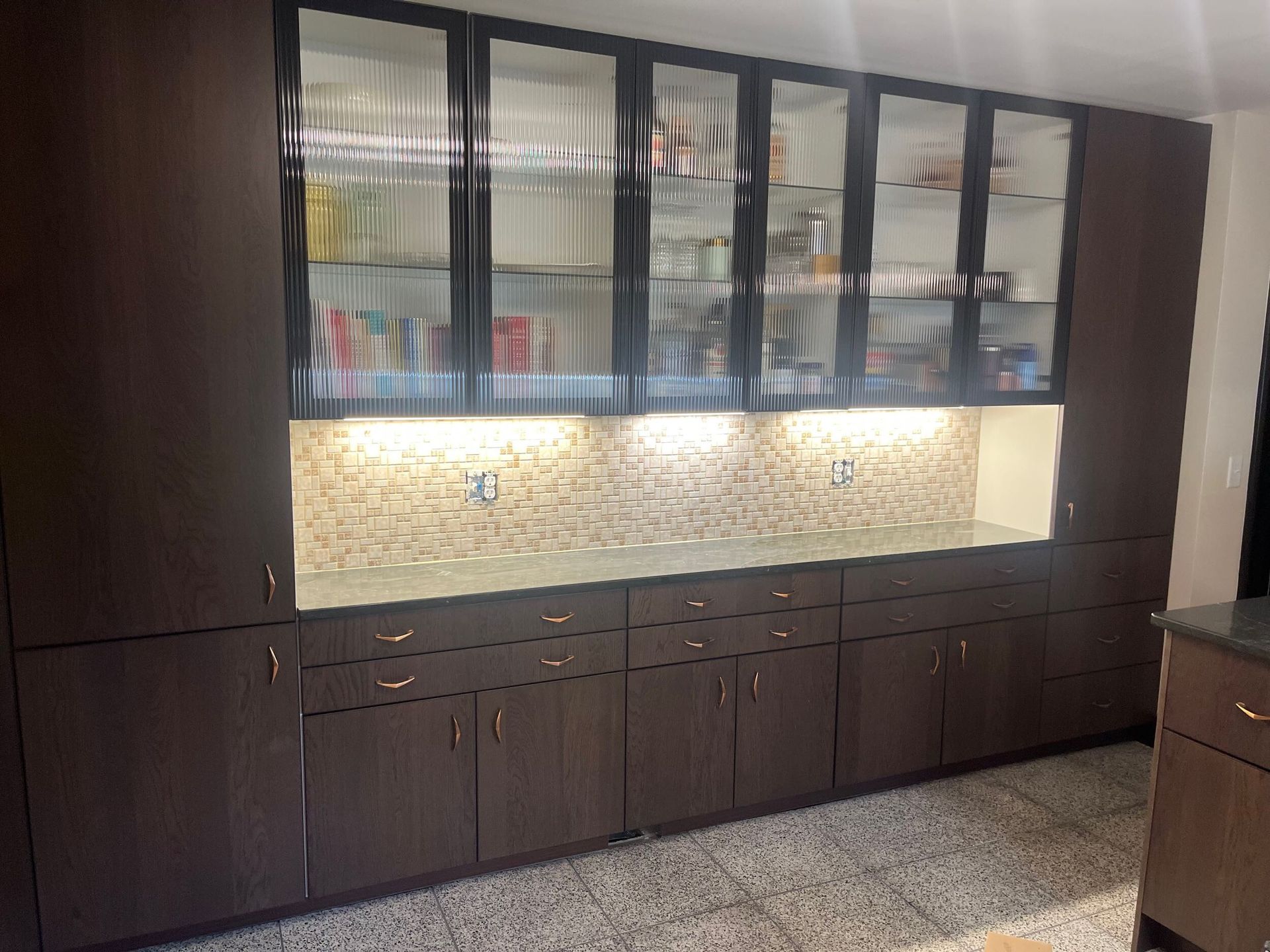 Dark wood kitchen cabinets with glass fronts above a beige backsplash and speckled countertop.