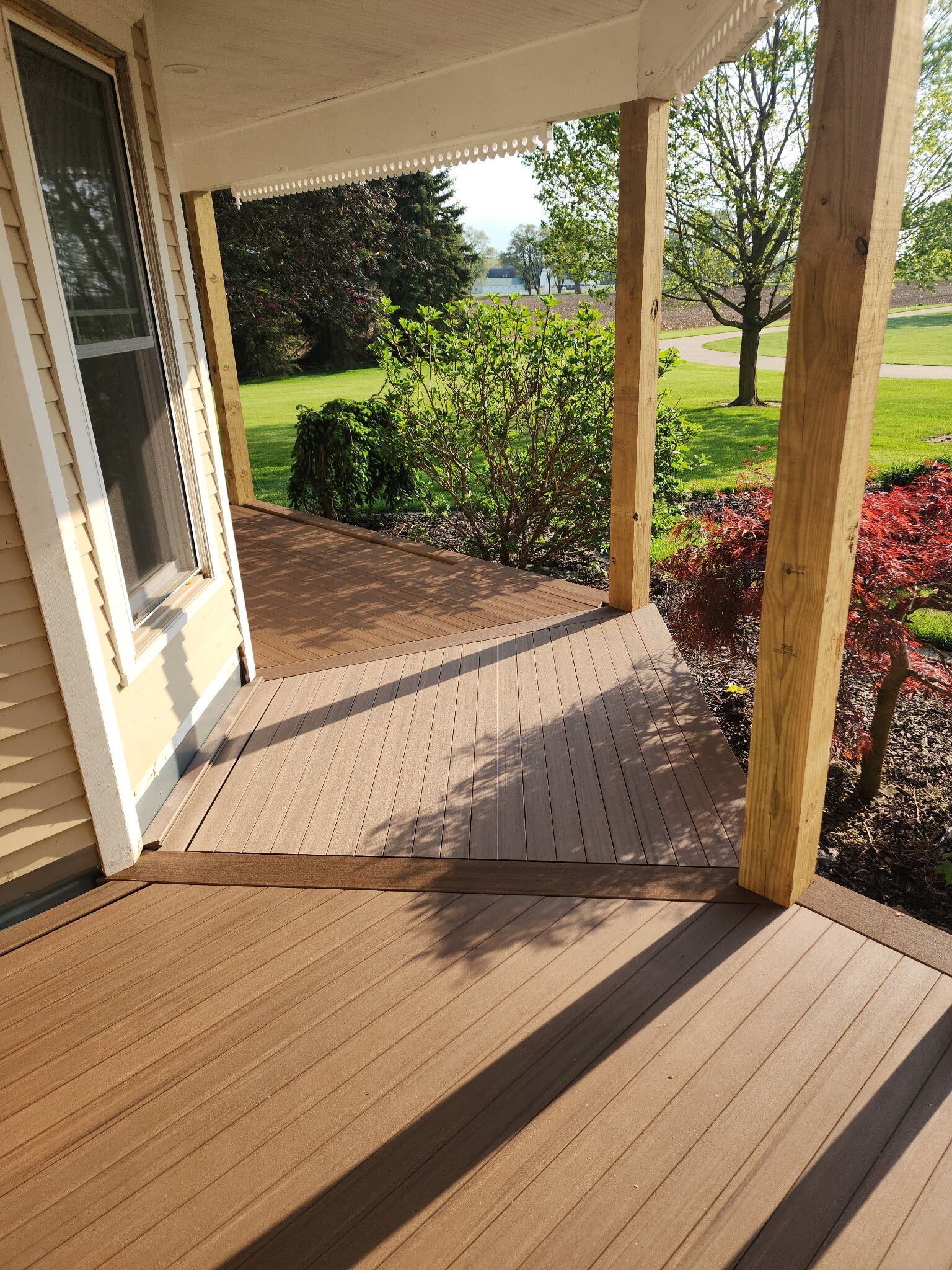 Front porch with tan floor, wooden posts, and a view of green shrubs and lawn in sunlight