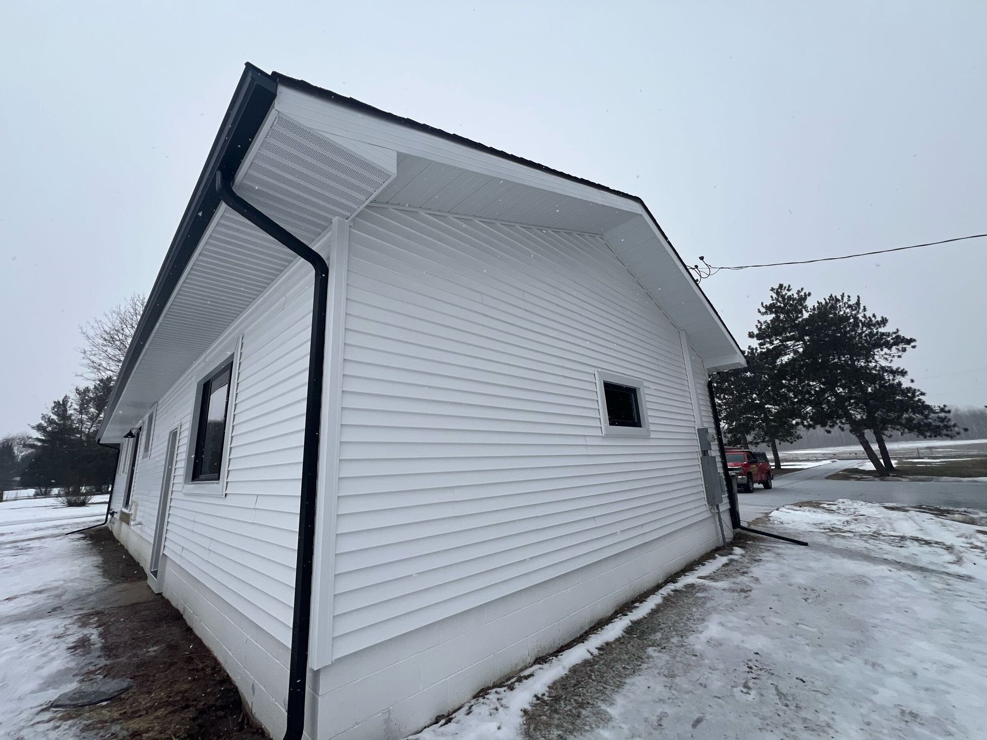 Snow-covered white house with black trim and siding, viewed from the side on a winter day.