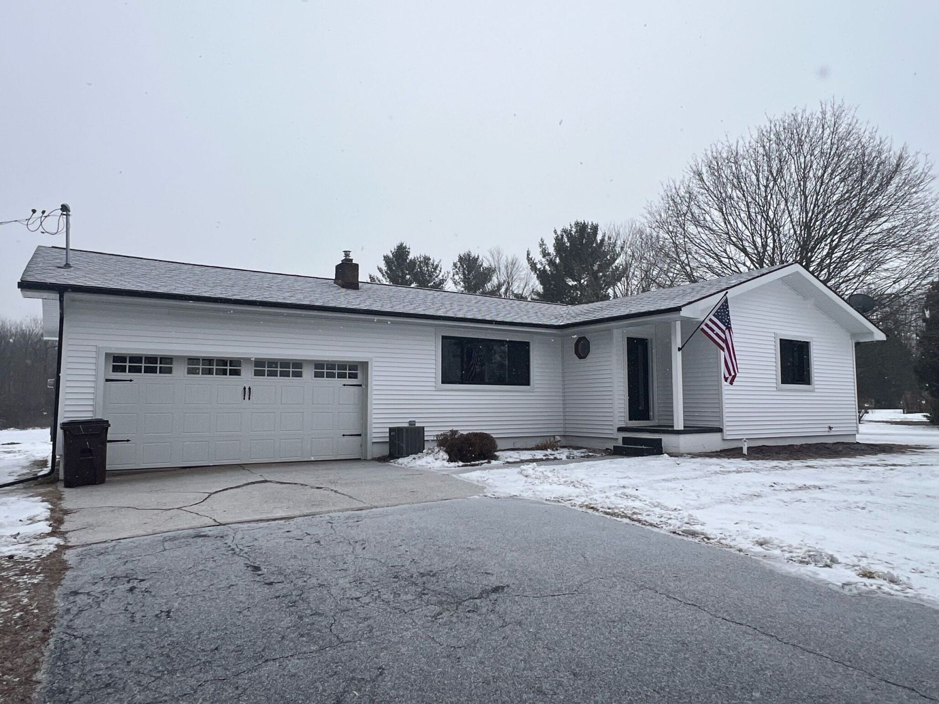 White house with a garage and American flag, driveway covered in snow.