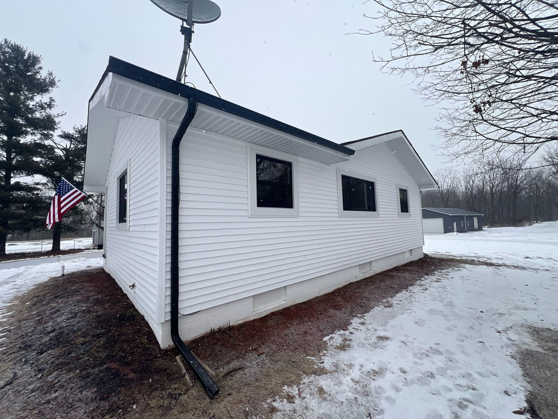 White house with black-trim windows and satellite dish, seen on a snowy yard on an overcast day