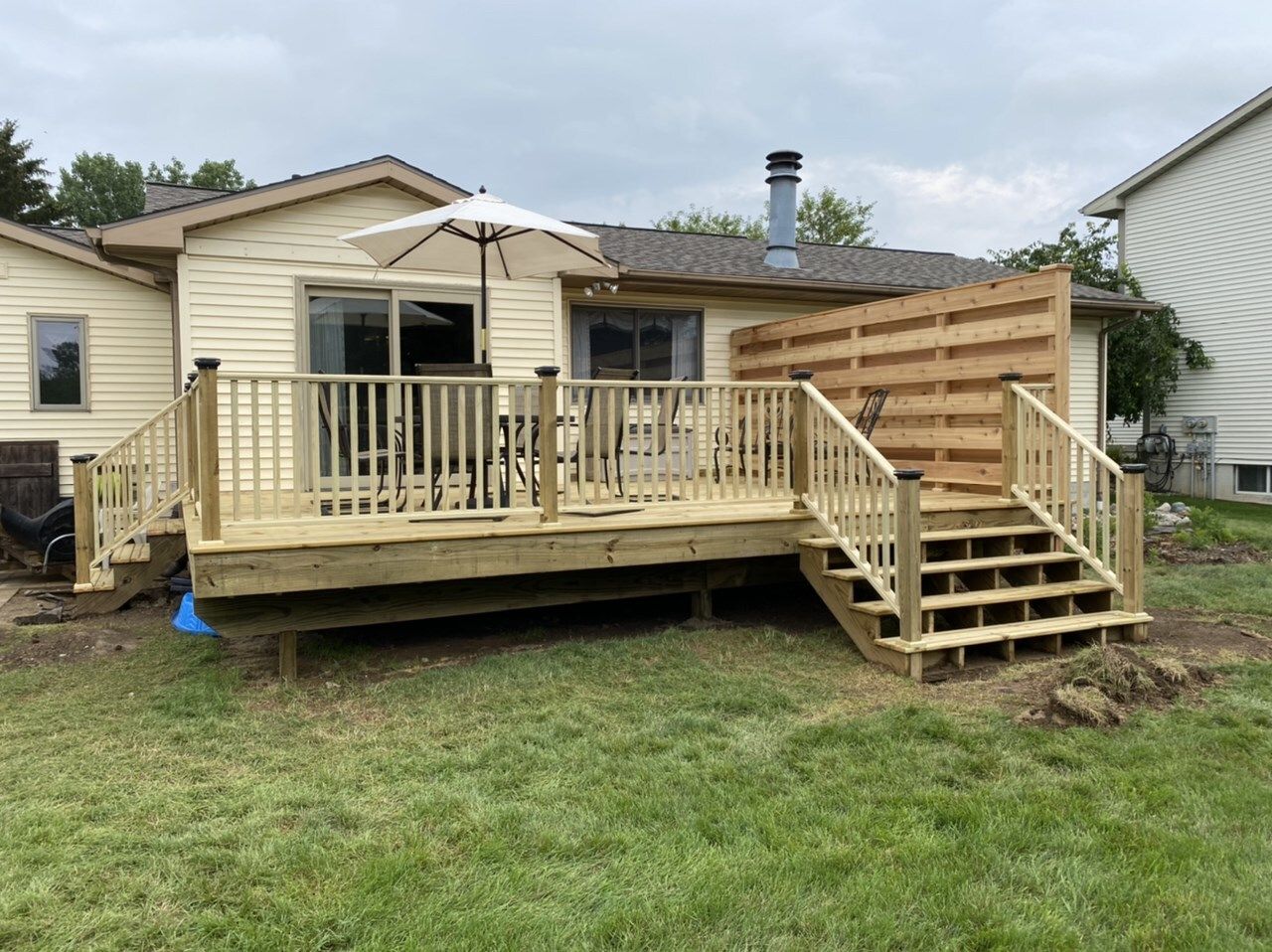 Wooden deck with stairs attached to a beige house, with a patio umbrella and grassy yard.