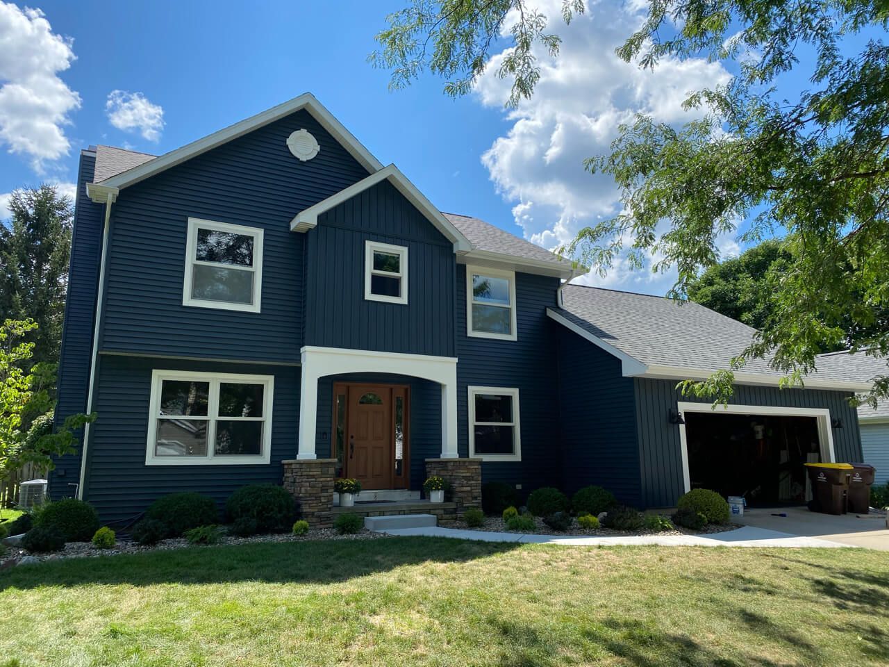 Blue-sided two-story house with white trim, brown door, and attached garage. Sunny day with blue sky and some clouds.