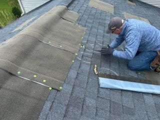 Worker kneeling on a shingled roof, repairing a section near a blue gutter guard.