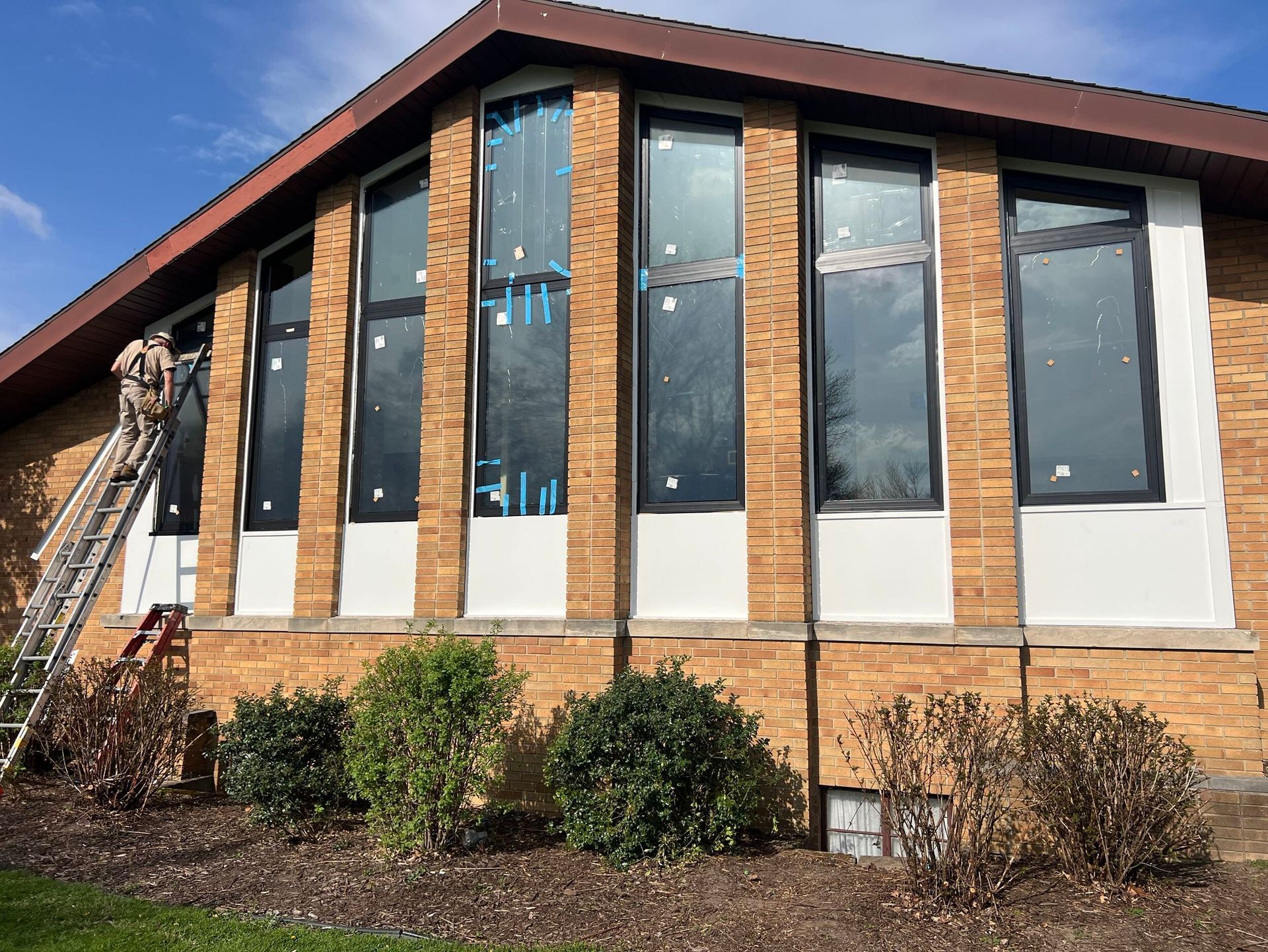 Brick building with tall dark windows, white lower panels, and shrubs along the front.