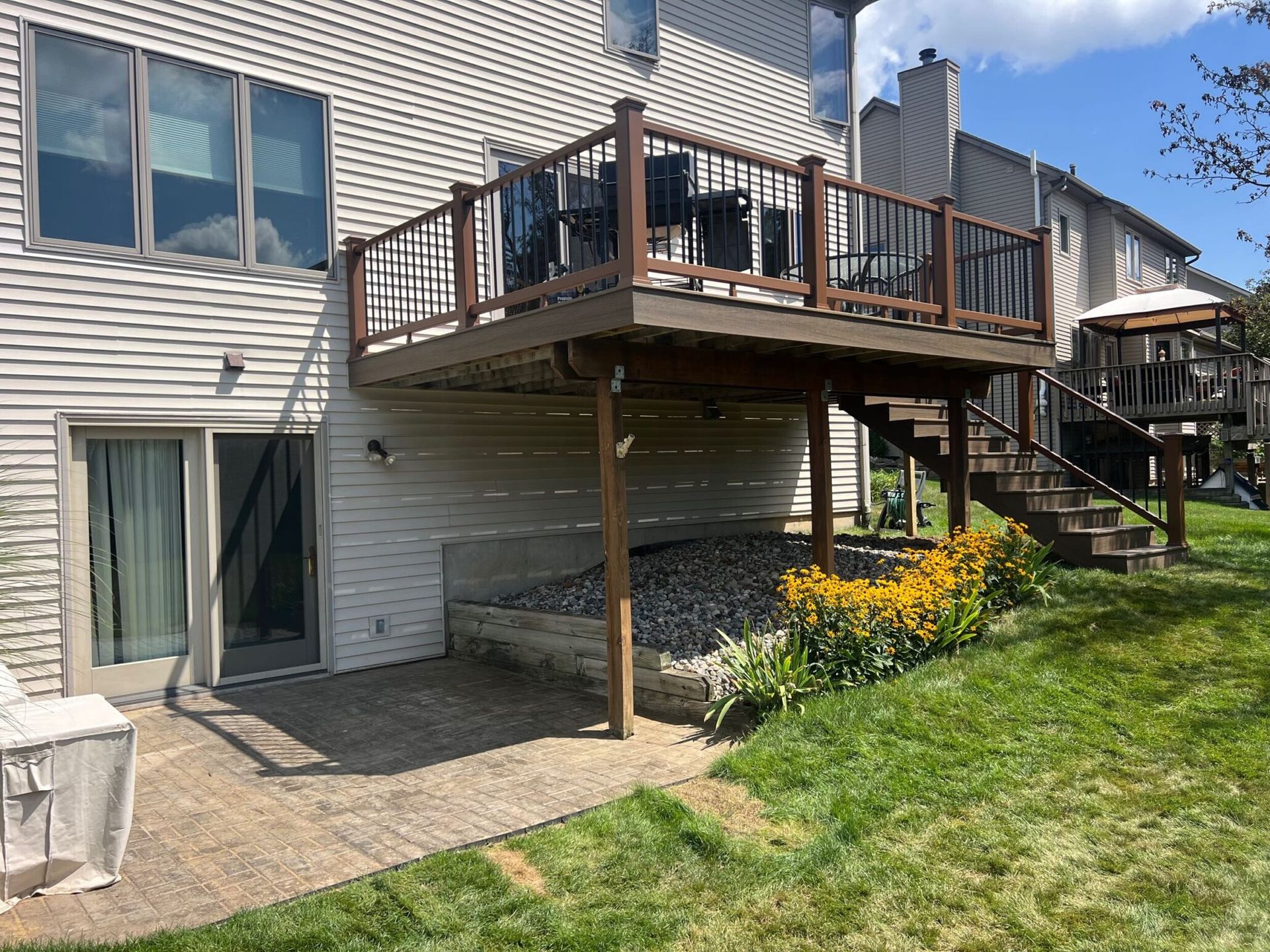 Backyard with a raised wooden deck, stairs, and patio next to a house with windows and a door.