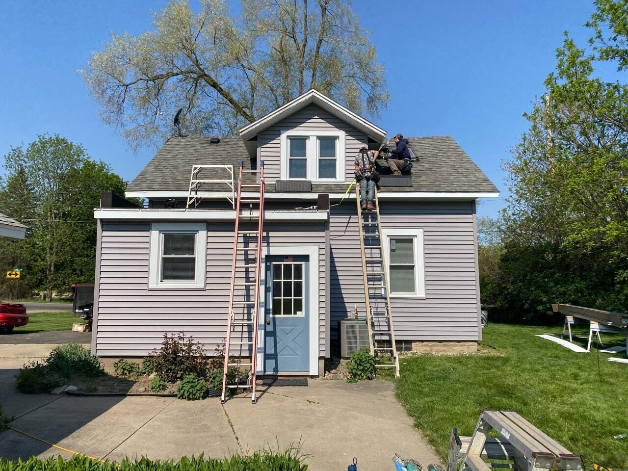 Gray house with two ladders leaning against the roof, viewed from the front in a sunny yard.