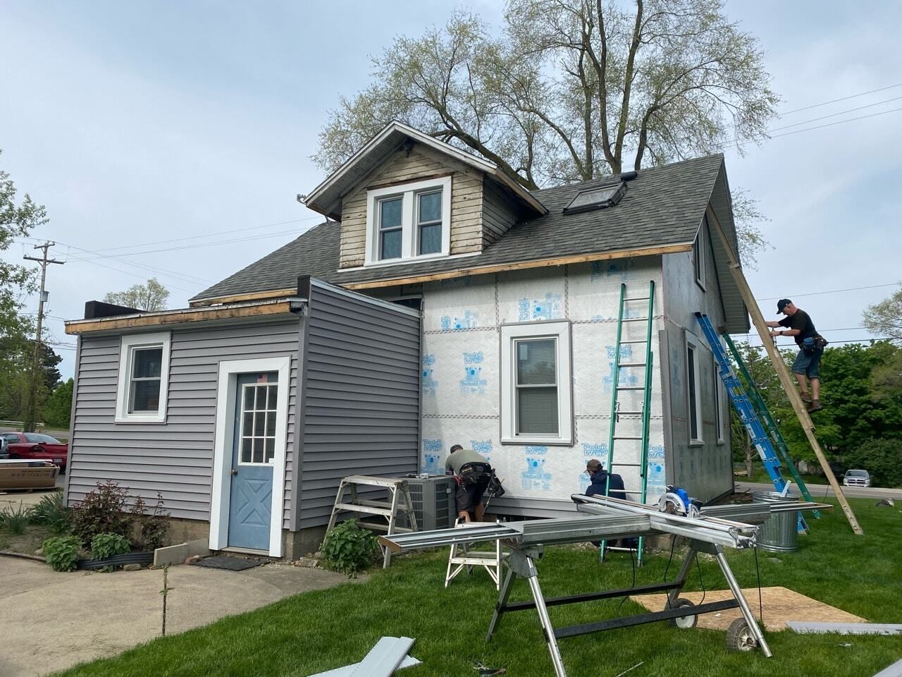 House under repair with siding partially removed, tools in front yard, and a person on a ladder at the side