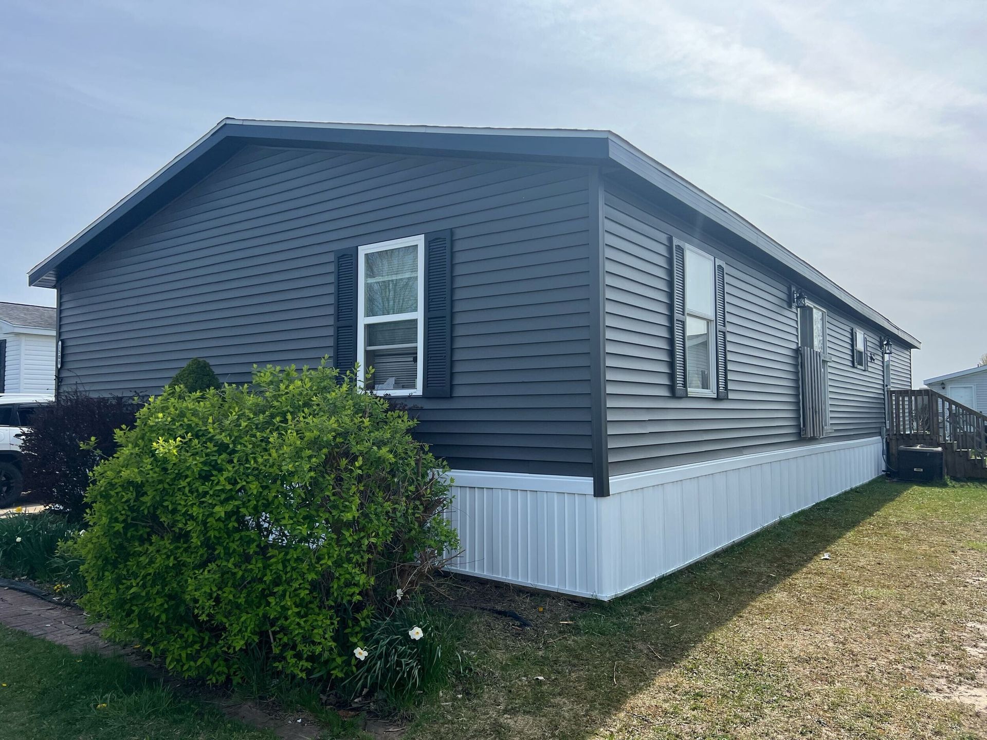 Gray manufactured home with white skirting and siding, viewed from the side beside a bush on a grassy lot.