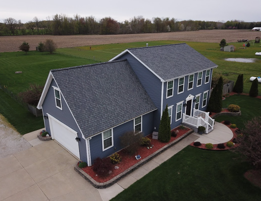 Blue two-story house with gray roof and white trim, seen from above in a rural setting with a driveway and lawn.