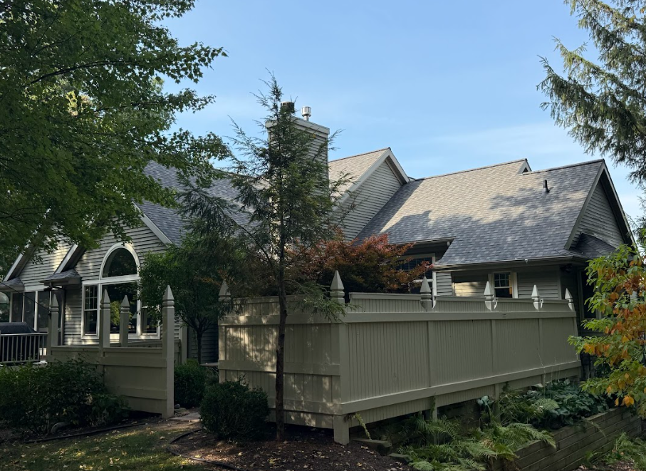 White house with steep gray roof behind a fenced porch, surrounded by trees and greenery.