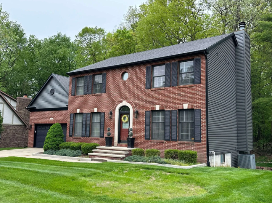 Two-story brick house with black shutters, a front porch, and a single-car garage on a green lawn.