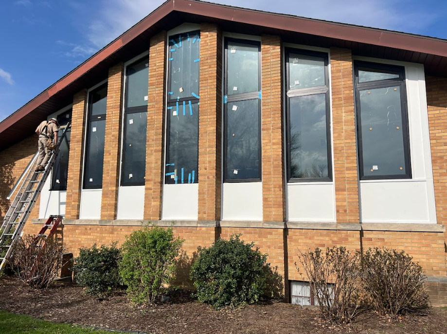 Building exterior with tall windows; person on ladder working on the windows. Brick wall, blue sky.