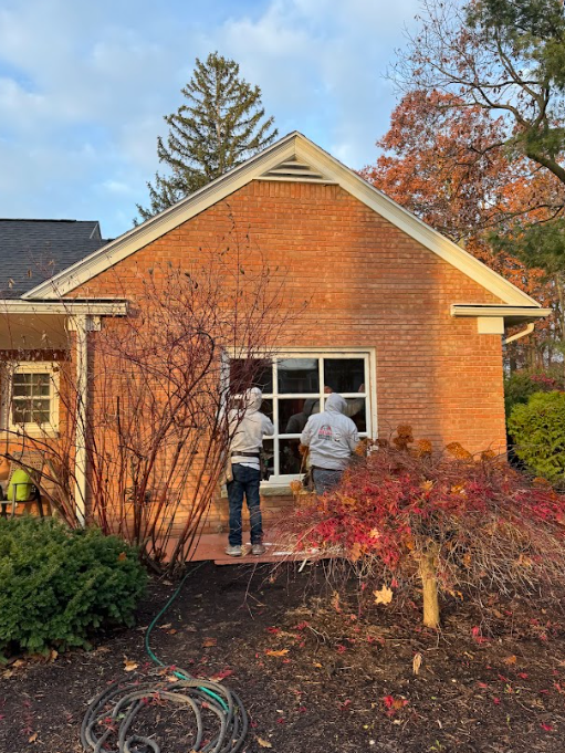 Two workers installing a window on a brick building with fall foliage in the yard.