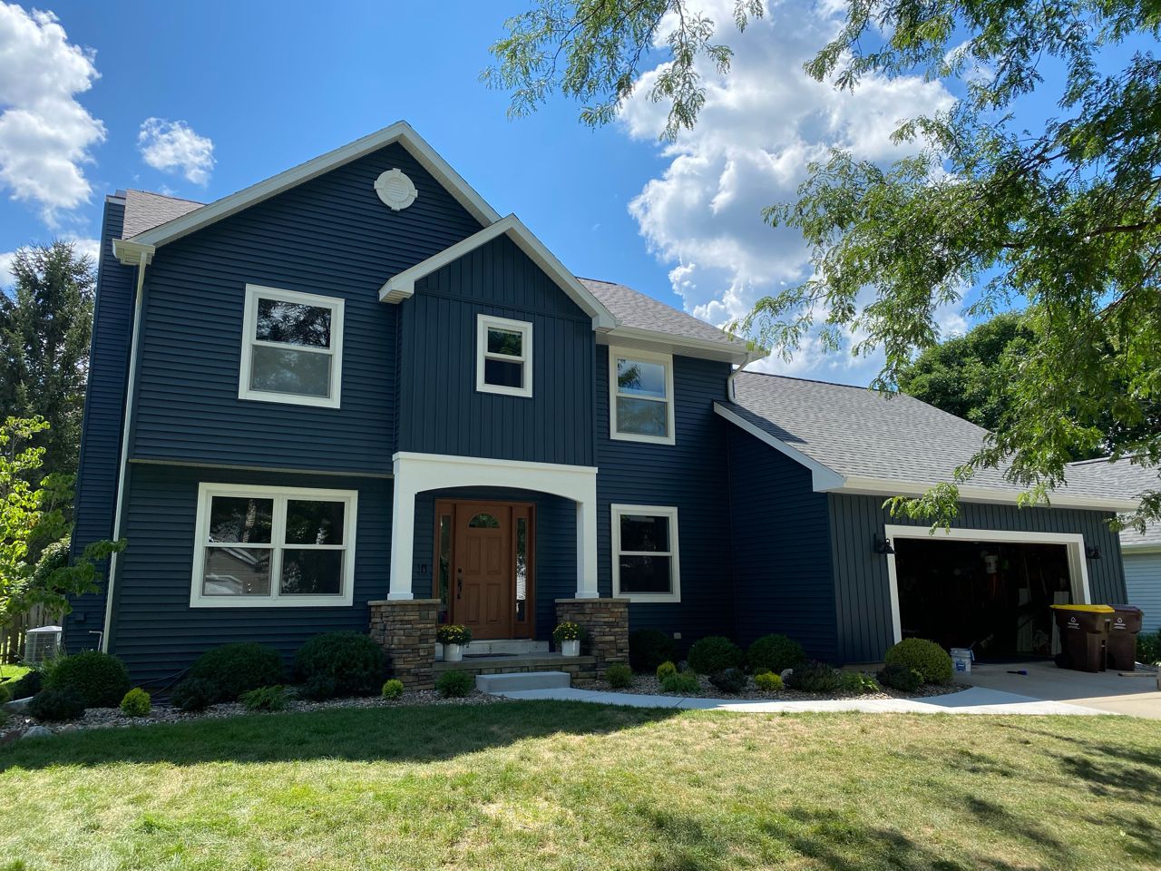 Blue two-story house with white trim, front porch, and attached carport under a sunny sky