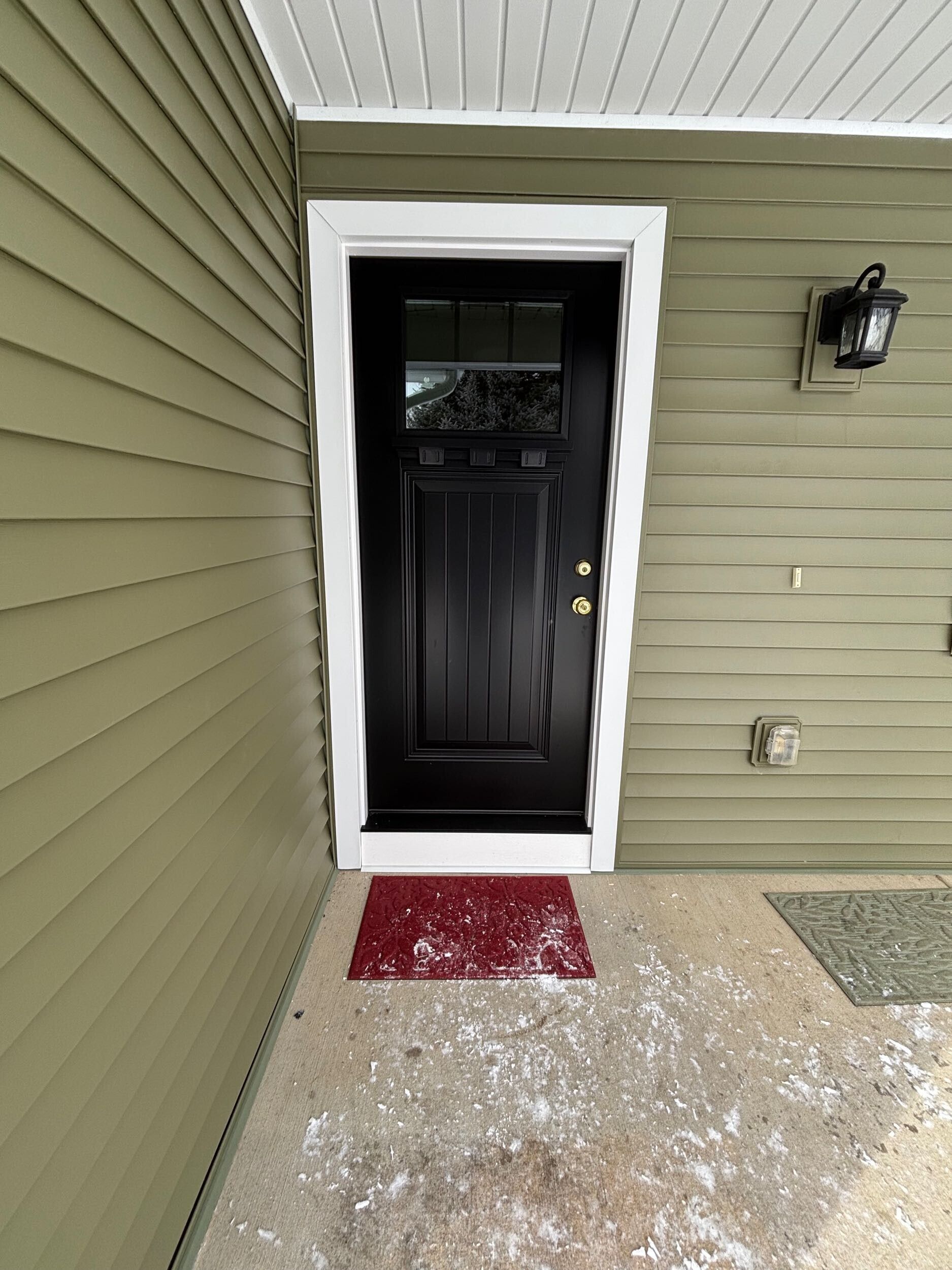Front entrance with black door, white trim, red doormat, and a wall light on beige siding.