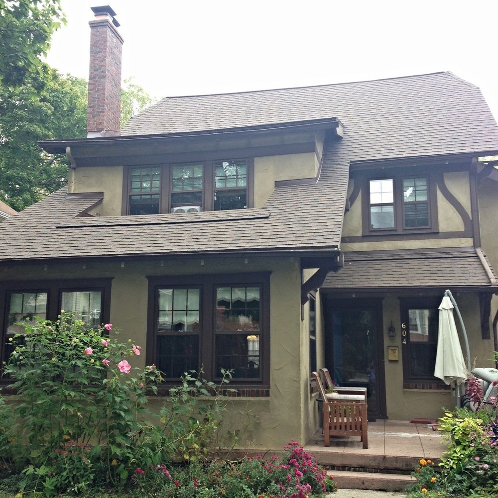 Two-story brown house with a chimney, front porch, and garden greenery.