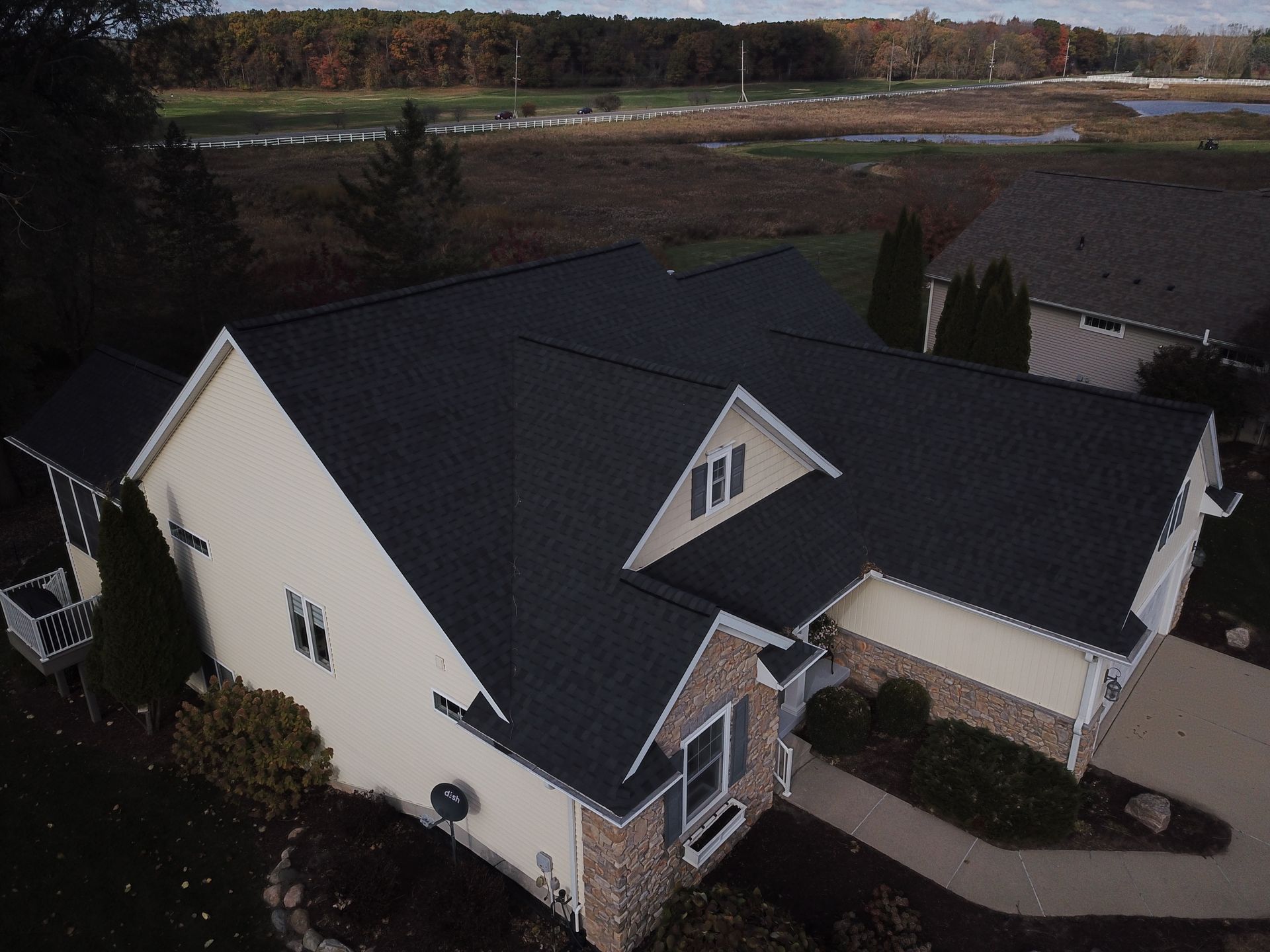 Aerial view of a beige house with a dark roof and curved walkway, surrounded by trees and lawn.