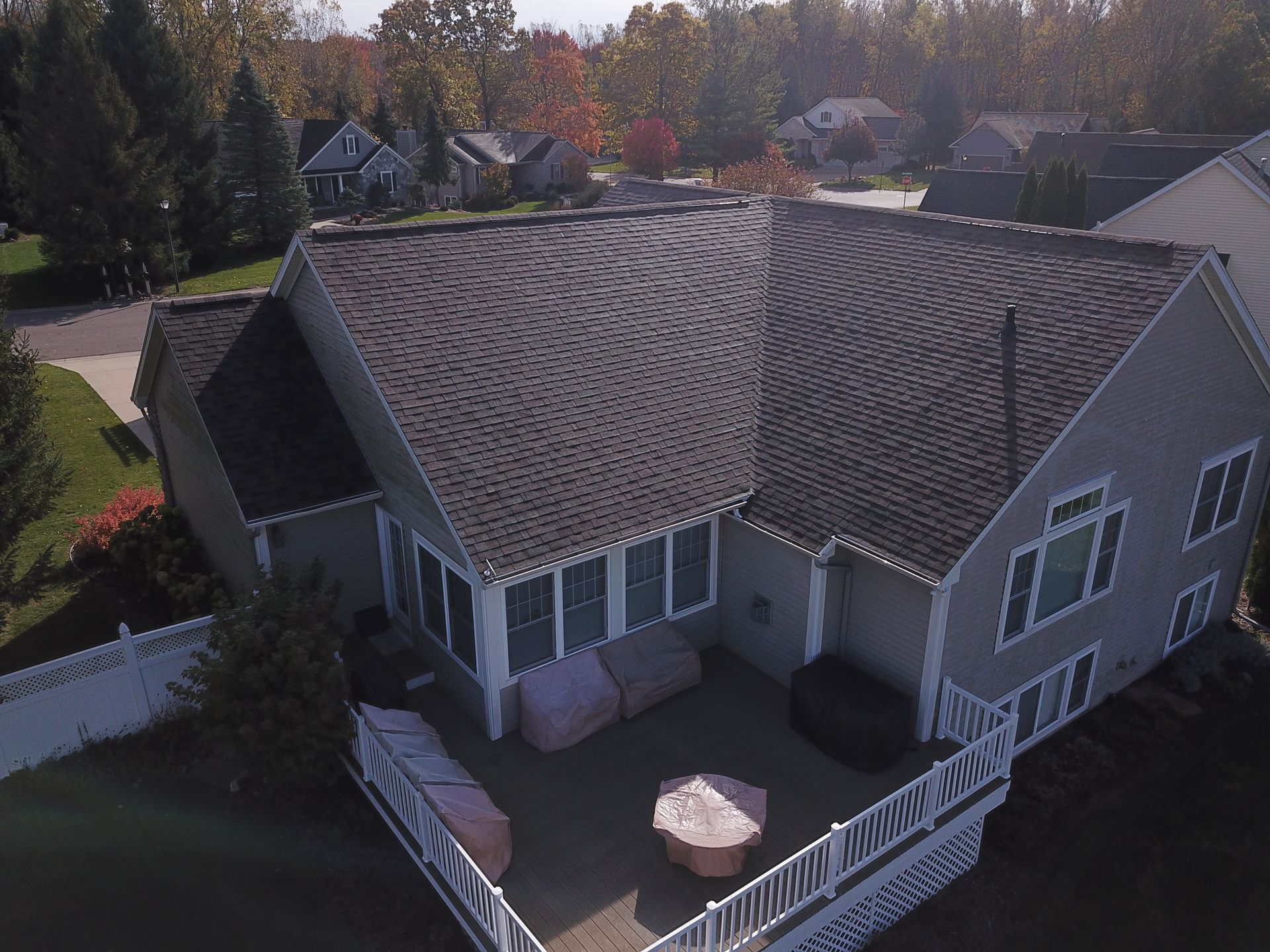 Aerial view of a gray house with a large roof, white deck, and patio furniture surrounded by trees.
