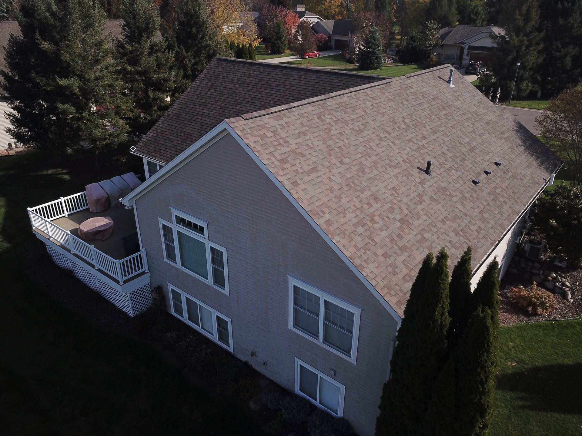 Aerial view of a beige house with a gray roof and white deck, surrounded by trees and greenery.