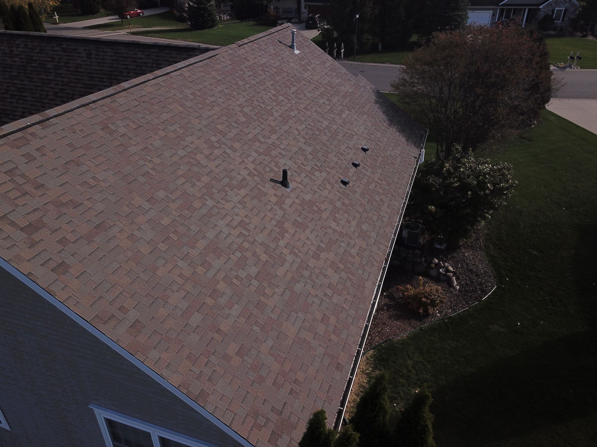 Aerial view of a shingled roof beside a lawn and shrubs, with several roof vents visible.