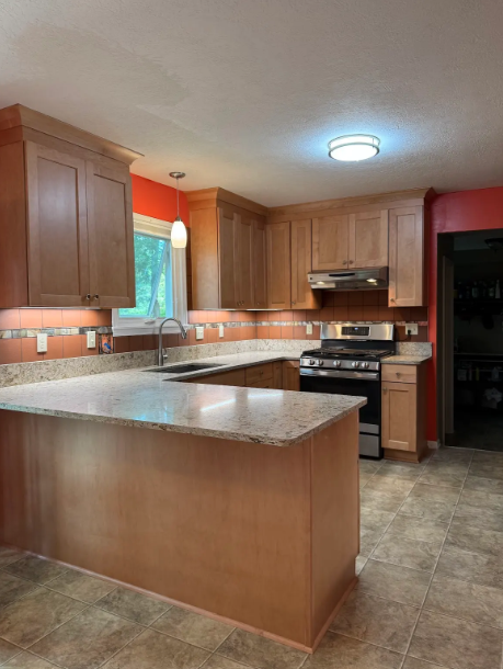 Wooden kitchen with granite island, stainless stove, and window with orange curtains.