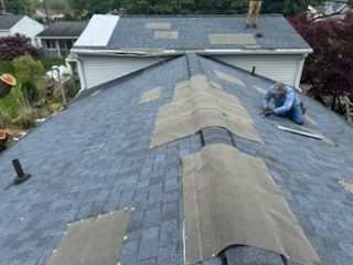 Worker repairing shingles on a steep gray roof near the ridge line