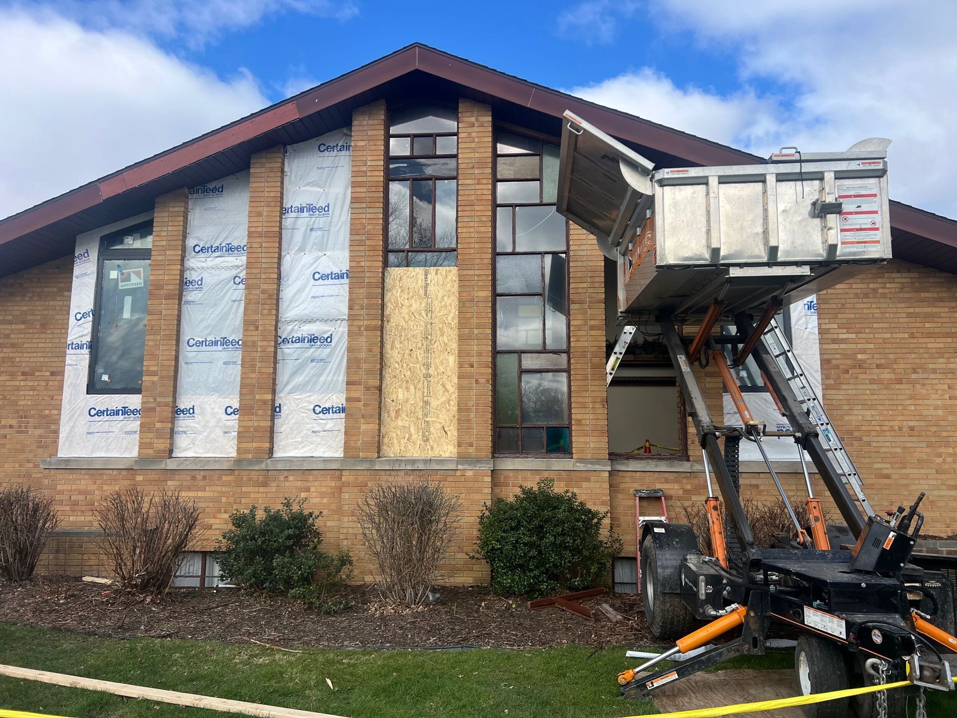 Workers in a boom lift repairing a brick building with wrapped windows and a steep roof