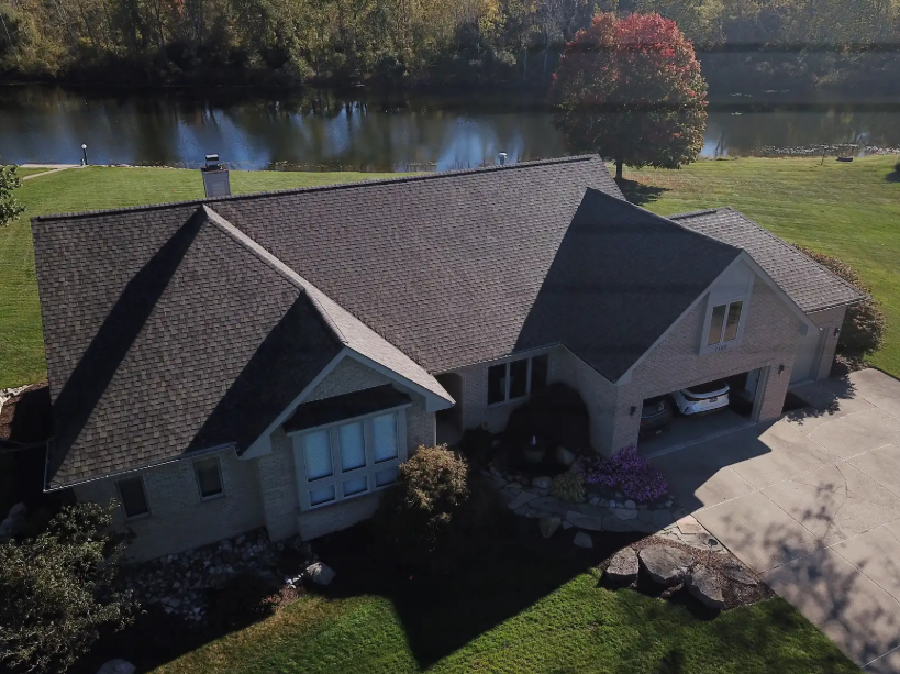 Aerial view of a large house with a dark roof beside a pond and trees