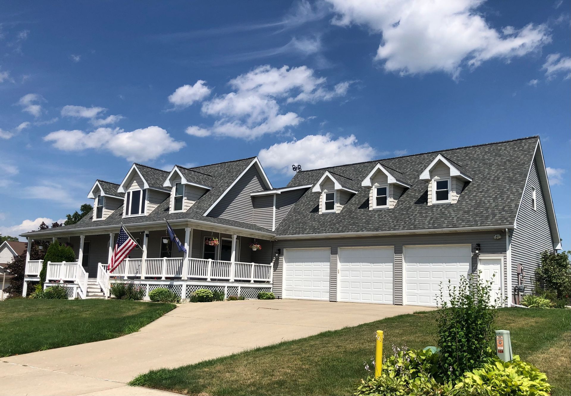 Large gray suburban house with a front porch, three white garage doors, and an American flag under a blue sky