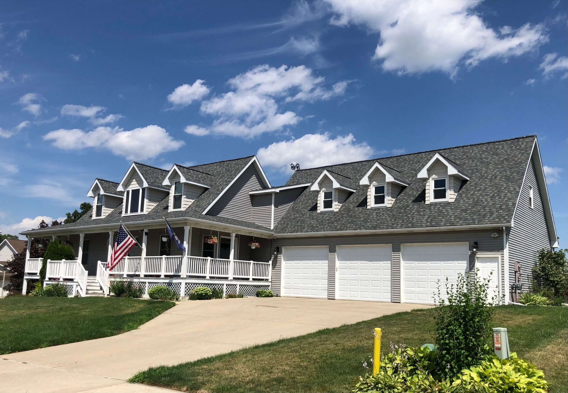 Large gray house with dormer windows, white garage doors, and an American flag under a blue sky