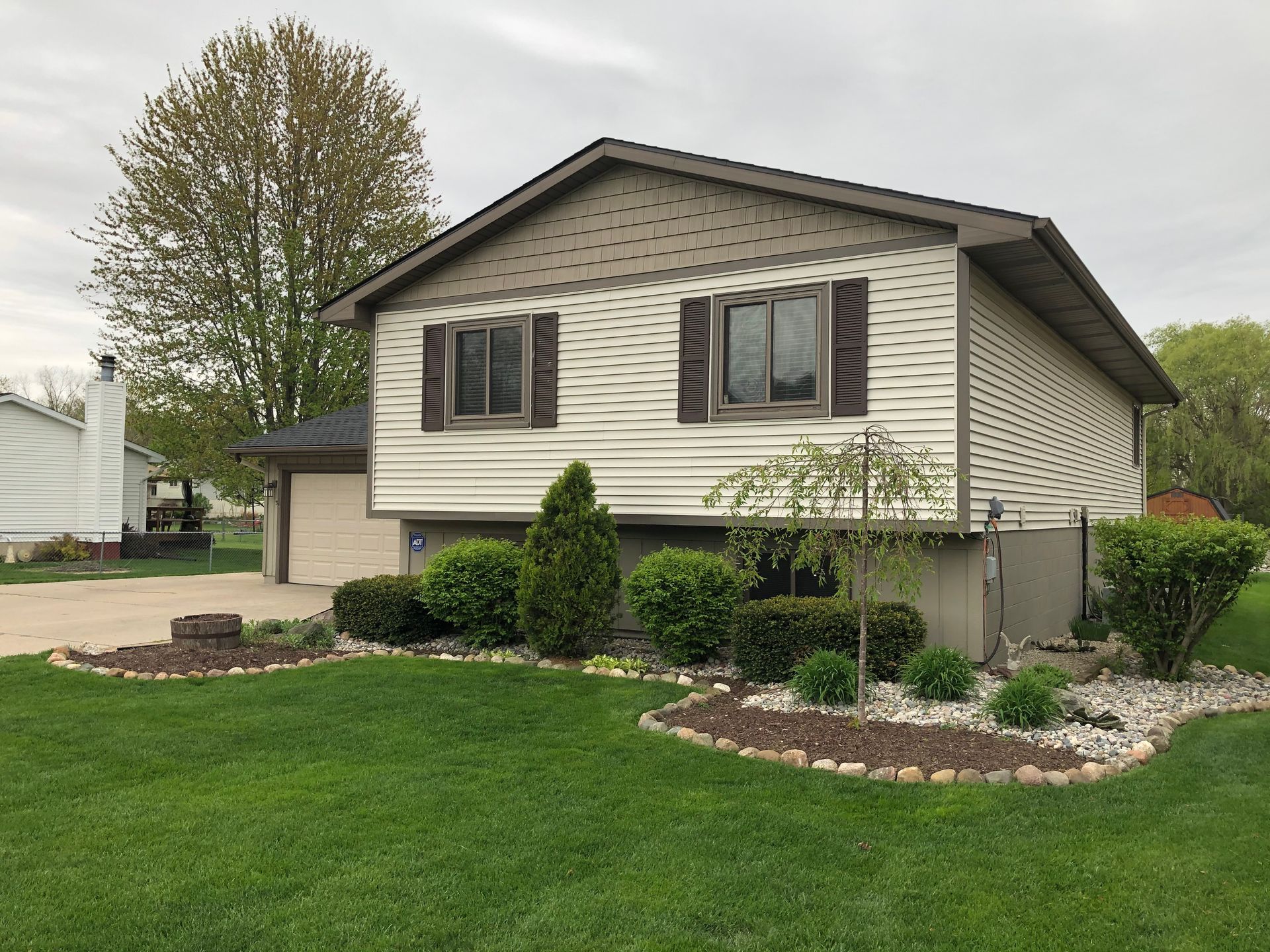 Suburban split-level house with beige siding, brown shutters, and a neatly landscaped front yard
