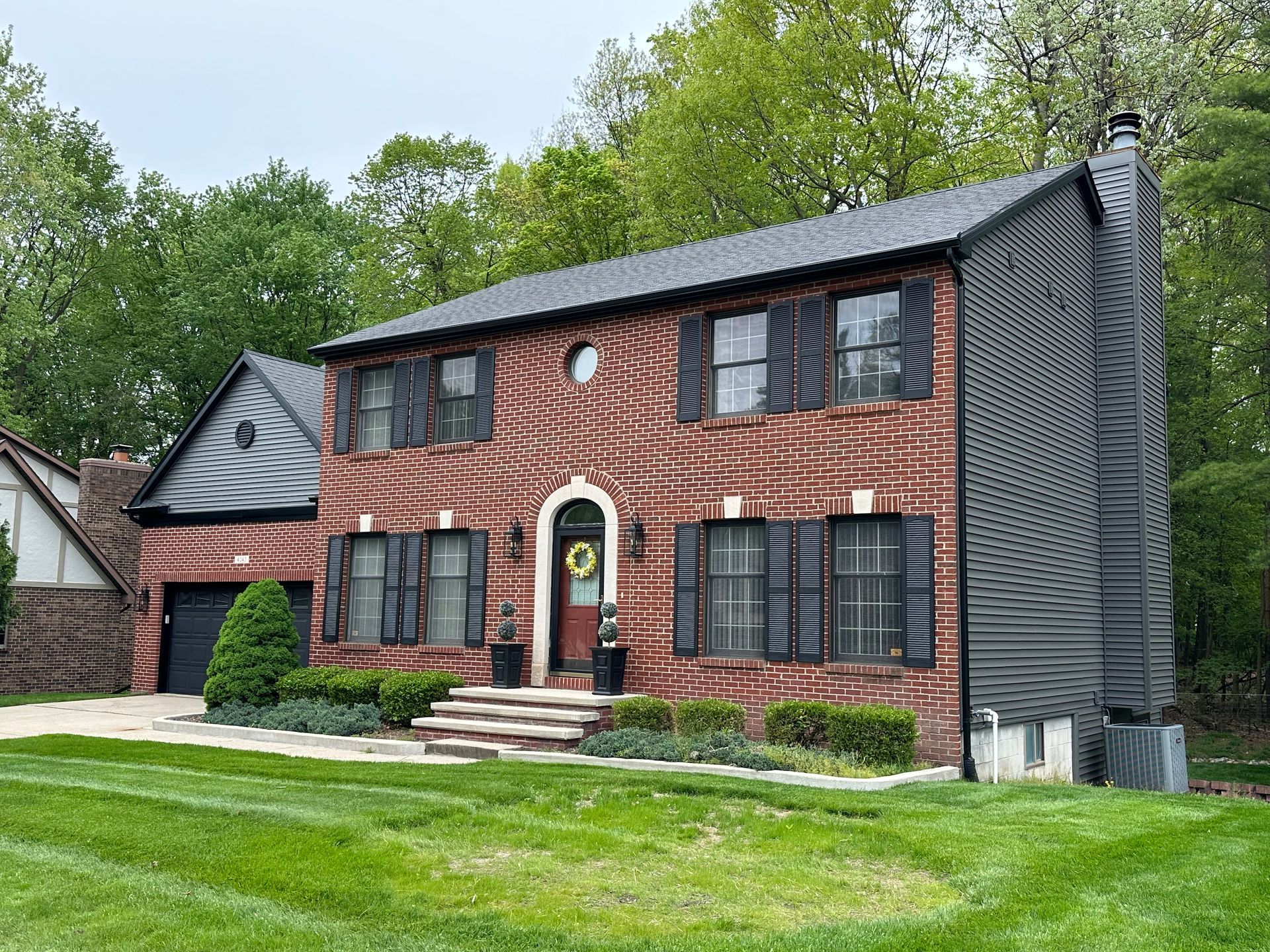 Two-story red brick house with black shutters, garage, and manicured lawn under trees