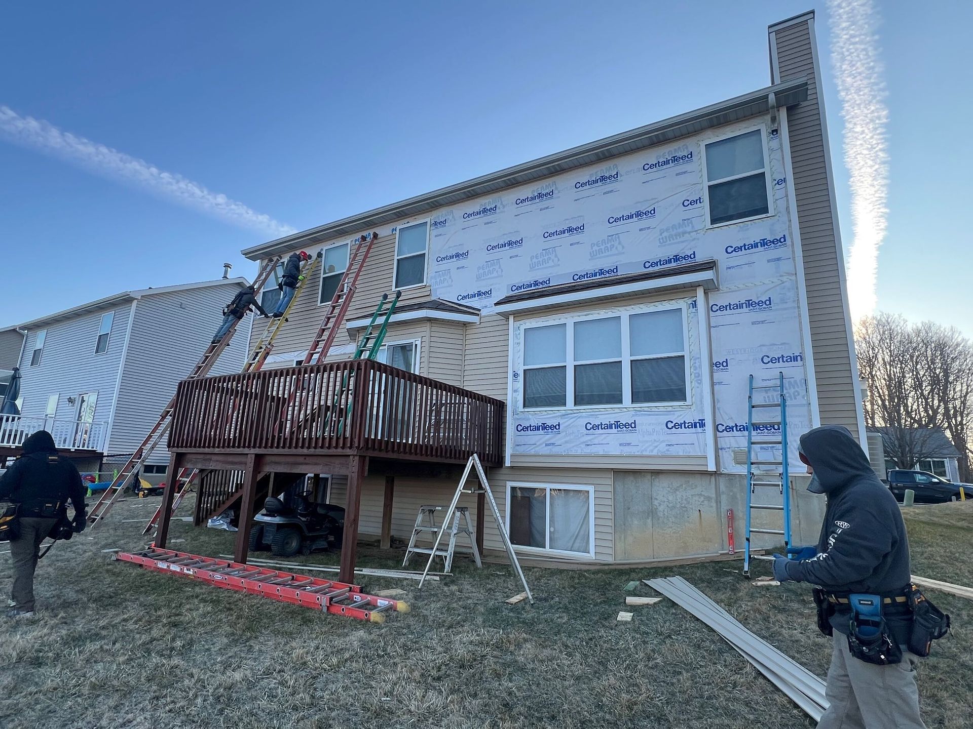 Workers install siding on a house with a raised deck and ladder in a frosty yard.