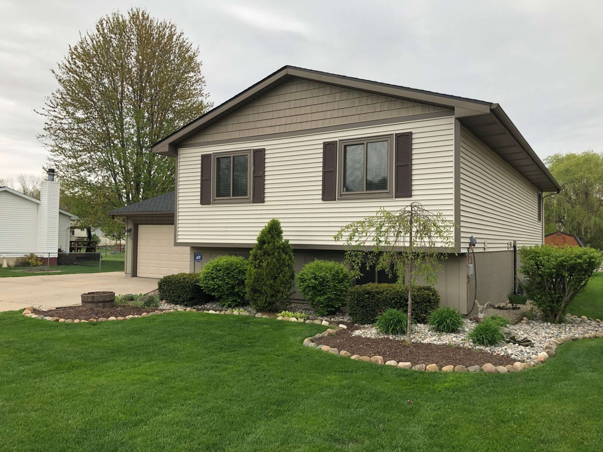 Suburban split-level house with beige siding, brown shutters, and a landscaped front lawn.