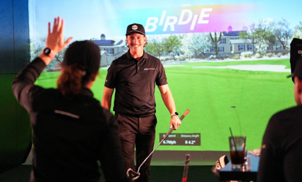 Man standing in a golf simulator studio waving, with virtual golf course projected behind him.