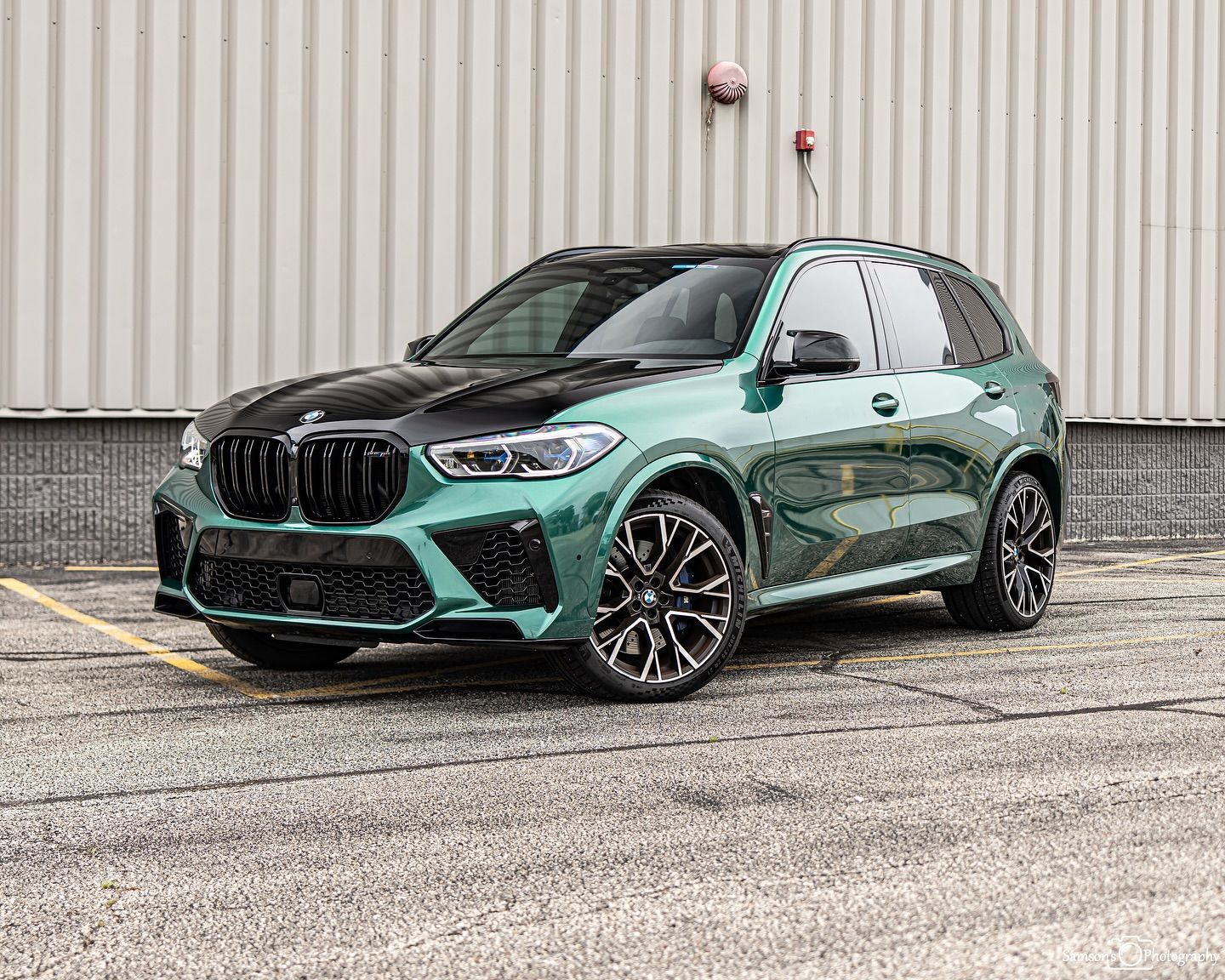 Green and black BMW X5 M SUV parked on asphalt. Black hood, gray building background.
