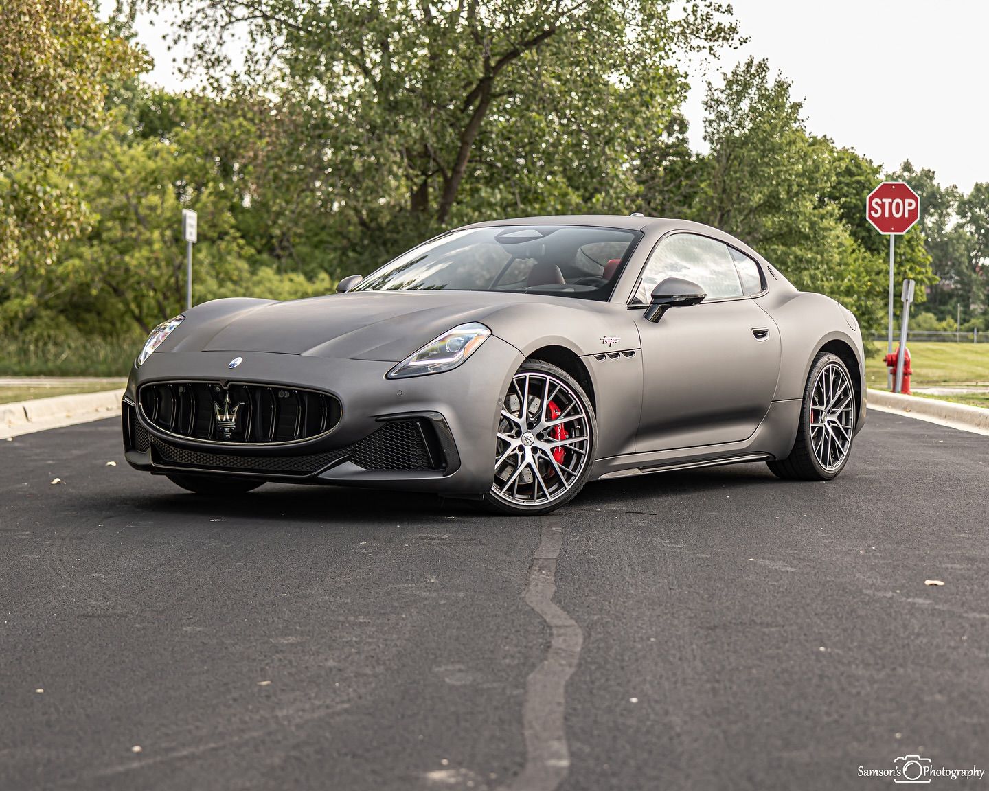 Matte gray Maserati sports car parked on asphalt road near trees and a stop sign.