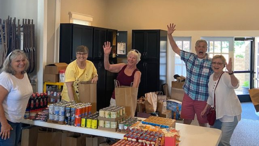 Five people wave near a table with canned goods and drinks inside a room.