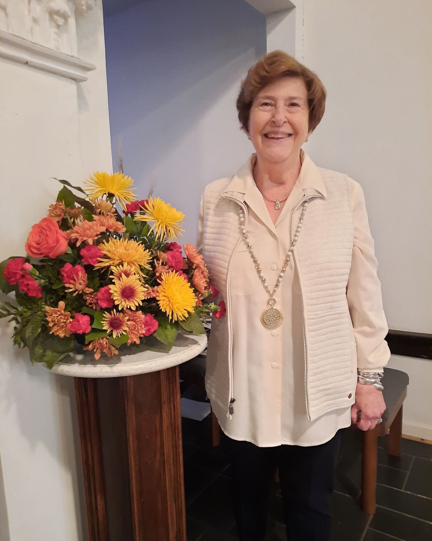 Woman in cream-colored outfit smiles next to a flower arrangement on a wooden pedestal.