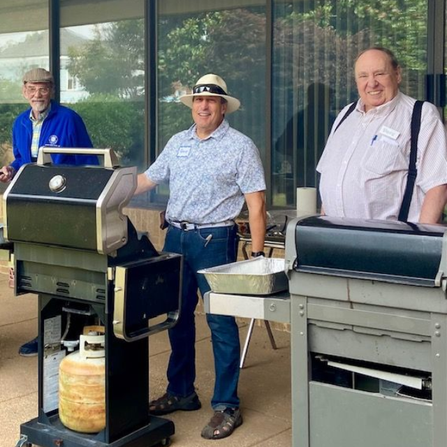 Three men grilling outside: one in a hat tends a grill, others watch.