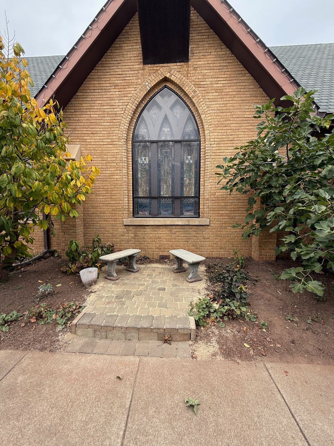 Church entrance with stone facade, arched window, two benches, and brick walkway. Autumn foliage.