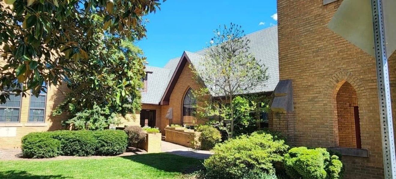 A beige brick church with a gray roof, surrounded by green trees and bushes, on a sunny day.