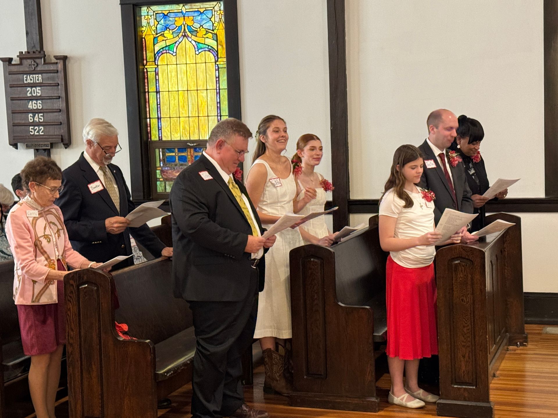 People singing in a church, holding papers. Stained glass window in background. Dark wood pews.