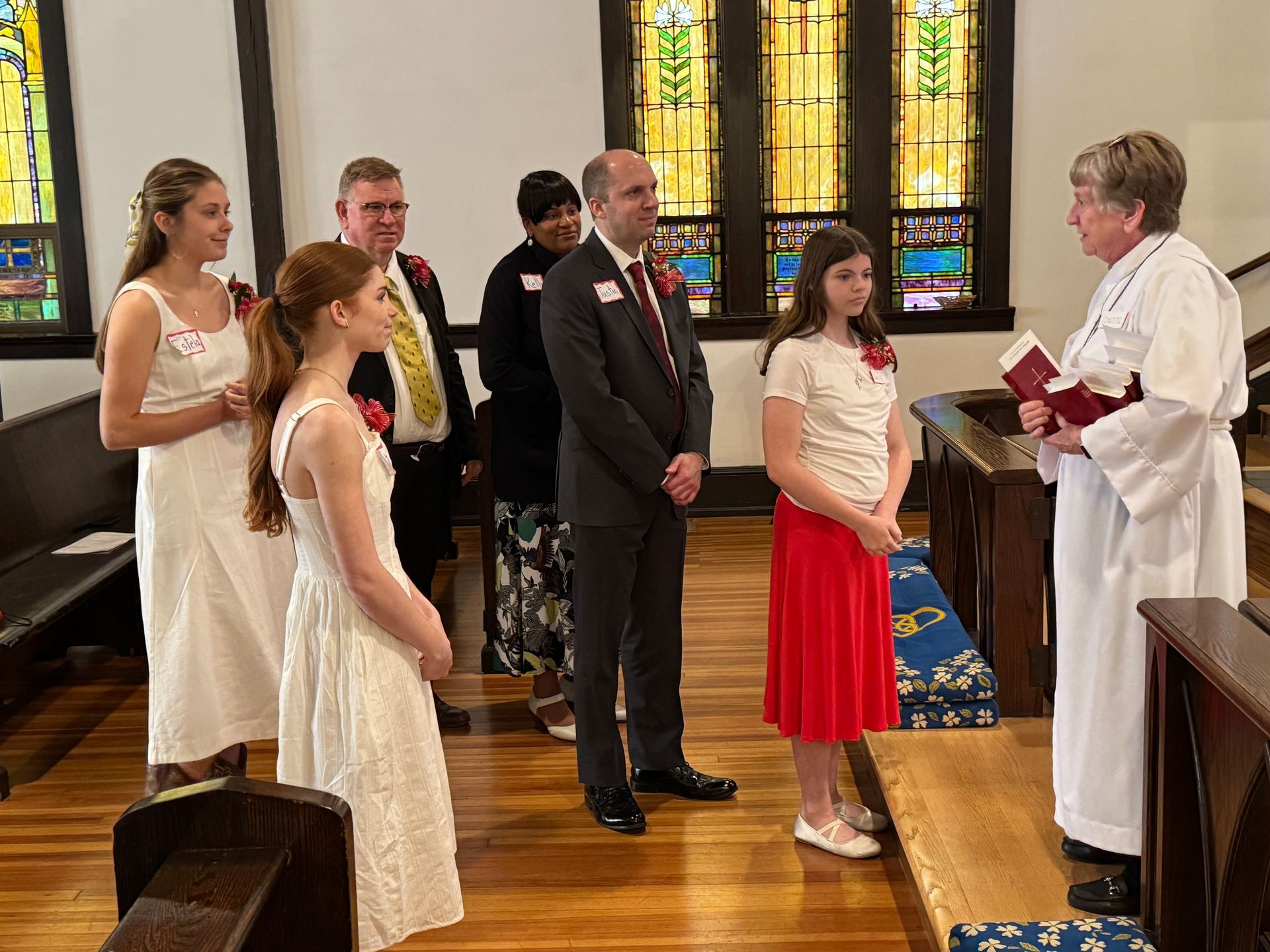 People in a church, standing before a person in white robes holding a book. Stained glass windows visible.
