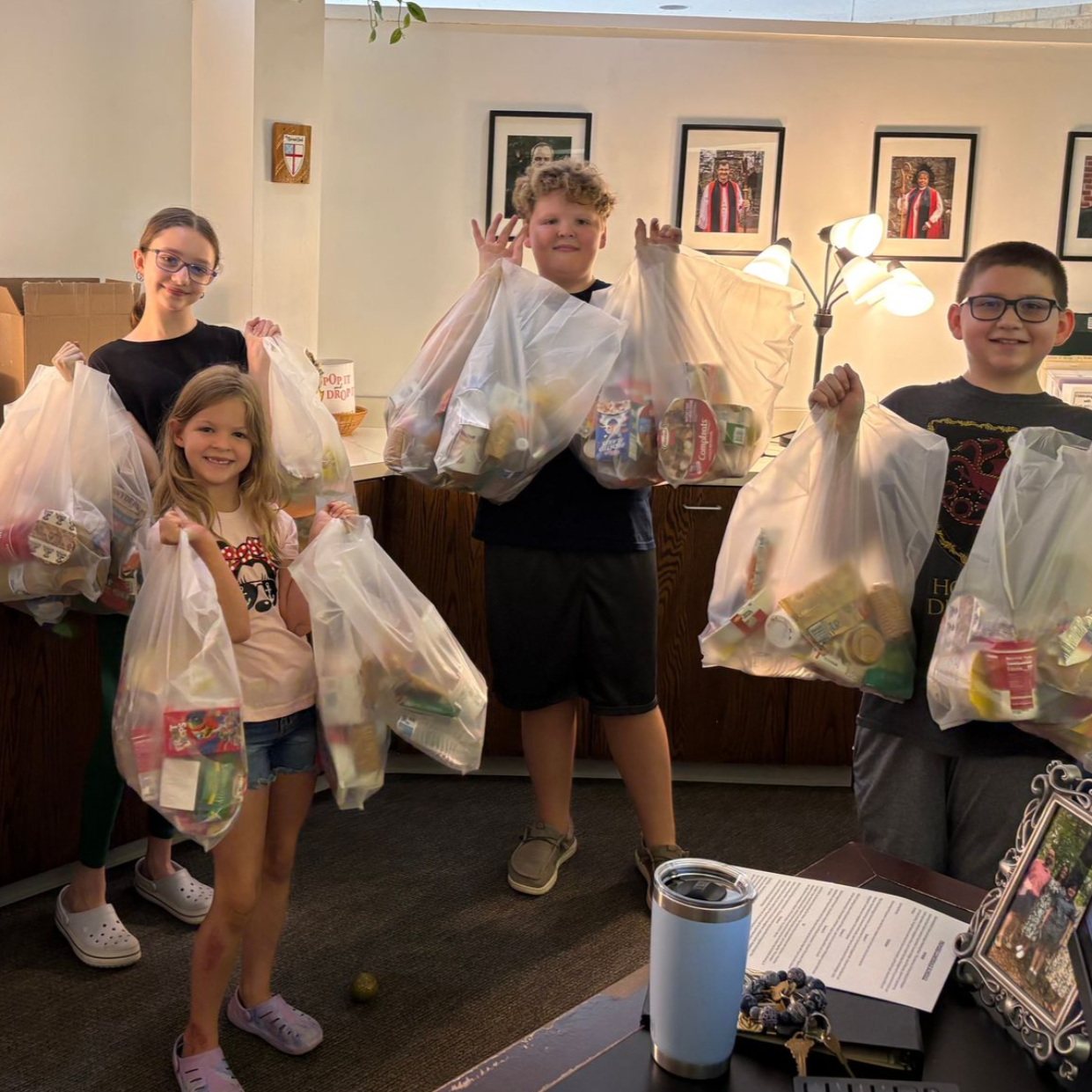 Four children holding bags of food, possibly for donation. They are smiling in an office-like setting.