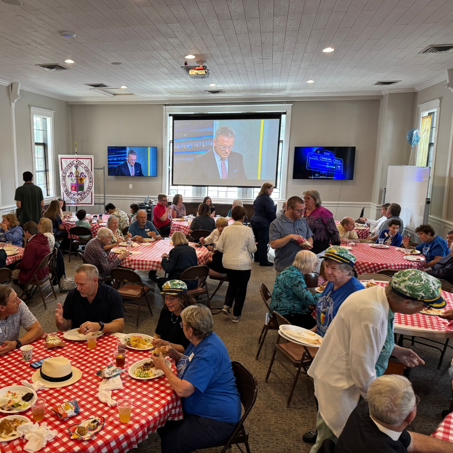 People at tables eating, watching screens in a room with red and white checkered tablecloths.